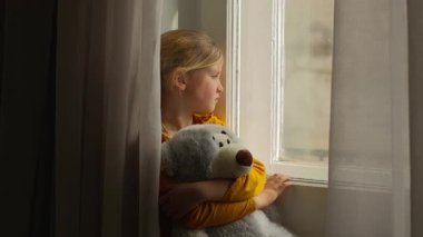 Hüsrana uğramış Kafkas kız çocuk yetim kız elinde oyuncak ayıyla pencereden bakan kız çocuğu sıkılmış kurban masumiyeti. Depresyonda. Evlatlık velayetini bekliyor.