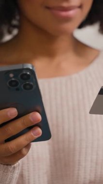 Cep telefonu ve banka kredi kartı alışverişi yapan mutlu, gülümseyen Afro-Amerikan kadın ev alışverişinde e-ticaret uygulaması internetten yemek siparişi veriyor.