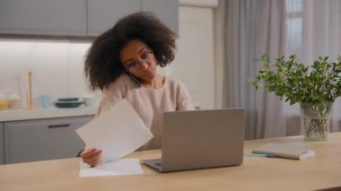 Cep telefonuyla konuşan çok görevli Afrikalı Amerikalı kadın ev işkadını bilgisayarını kullanarak evrakları yönetmeyi düşünüyor.