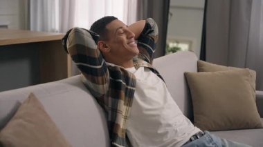 Gülümseyen, dinlenen, Afro-Amerikan erkek, etnik erkek. Klima içeride, kanepede dinleniyor. El ele tutuşup ev sahibinin arkasına yaslanıyor.