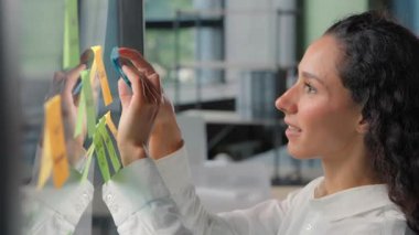 Gülümseyen mutlu İspanyol iş kadını ofis neşe dolu iş kadını kız şirketinin CEO 'su cam üzerine yapışkan notlar alıyor. Sonuç olarak pazarlama stratejisi çıkartmaları yaratıyor.