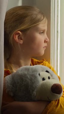 Üzgün, küçük, üzgün, beyaz çocuk yetim kız pencereden bakan, oyuncak ayı kucaklayan, ebeveyn evlat edinme velayetini bekleyen, evde hayal kırıklığı yaşayan, izolasyon karantinasından muzdarip çocuk.