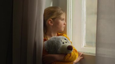 Üzgün, küçük, üzgün, beyaz çocuk yetim kız pencereden bakan, oyuncak ayı kucaklayan, ebeveyn evlat edinme velayetini bekleyen, evde hayal kırıklığı yaşayan, izolasyon karantinasından muzdarip çocuk.