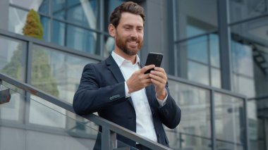 Başarılı gülümseyen orta yaşlı Kafkasyalı işadamı, akıllı telefon kullanarak, sosyal medya uygulamalarını kullanarak, e-posta sohbetlerini kontrol ederek, şehirli iş adamı liderinin cep telefonunu kaydırıyor.