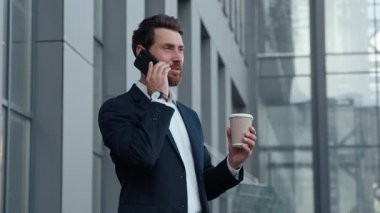 Heyecanlı gülümseyen mutlu Kafkasyalı iş adamı şehirde cep telefonuyla konuşuyor. Kaygısız bir iş adamı, elinde kahve, elinde içki, akıllı telefon, iyi haber yöneticisinin iletişimini tartışıyor.