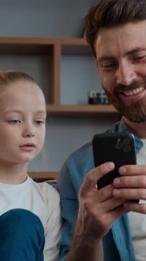 Oturma odasında mutlu bir aile babası, güzel bir kız çocuğuyla telefonda konuşuyor, mobil sosyal medya içeriği hakkında konuşuyor, evdeki kanepede dinleniyor, beyaz bir baba ve kız çocuğu online alışveriş yapıyor.