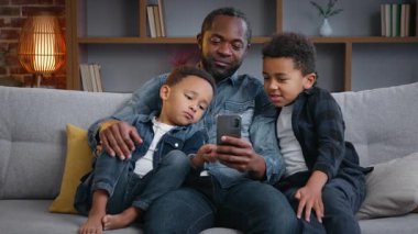 Mutlu Afro-Amerikan ailesi evde cep telefonu uygulaması kullanıyor. Babaları akıllı telefon aletlerine bakıyor. Gülümsüyor, sosyal medya çocuklarıyla konuşuyor.