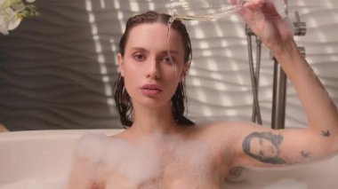 Beyaz kadın, sıcak köpüklü küvette dinleniyor şampanya dolduruyor suya alkol döküyor düşünceli bayan kız kameralı ev banyosu spa atmosferine bakıyor yıkanma havasında banyo yapıyor.