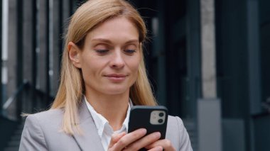 Gadget müptelası yetişkin beyaz iş kadını şehirde telefonla dolaşıyor odaklanmış iş kadını gülümsüyor akıllı telefon ekranlı mobil uygulama izliyor e-posta kontrolü yapıyor profesyonel bayan