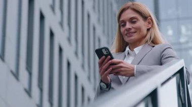 Ofis binasının yanındaki merdivenlerde telefonu olan orta yaşlı beyaz bir iş kadını şehrin merkezinde merdivenlerde durup gülümseyen mutlu iş kadını akıllı telefon kadınını kullanıyor.