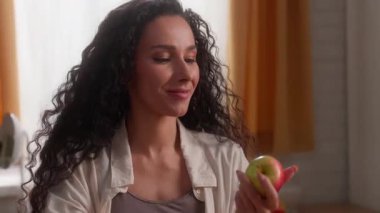 Mutlu İspanyol Latin kadın, hava mutfağına elma atıyor sağlıklı besleniyor, gülümsüyor, beyaz kadın, lezzetli meyveler yiyor, beslenme düzenliyor, kapalı alanda sağlıklı besleniyor, vitamin besleniyor.