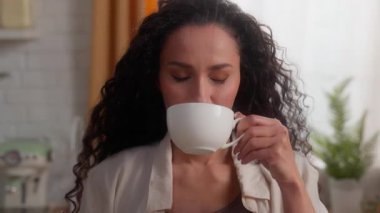 İspanyol beyaz kadın çay içiyor, göz kırpıyor, mutfağa gülümsüyor, Latin bayan kameraman kıza bakıyor, aromanın tadını çıkarıyor, sabah kahvaltısını hazırlıyor, sıcak bitkisel içeceği gevşetiyor.