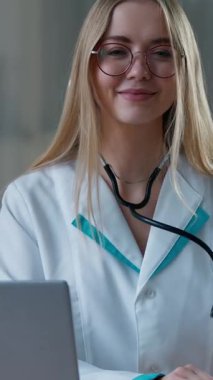 Klinikte poz veren beyaz kadın doktor masadaki steteskop tıbbi ekipmanlarını kameraya gülümseyen pediatrist kardiyologlara bakan dizüstü bilgisayarcı hemşire ile birlikte gösteriyor.