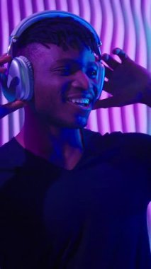 Neşeli duygusal Afrikalı Amerikalı adam fütürist neon klübünde kulaklıkla müzik karışımı dinliyor mutlu DJ erkek adam müzik dinliyor fütürizm alanında gülücük dansı yapıyor eğleniyor