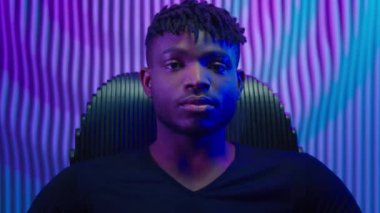 Neon karanlık stüdyosunda oturan etnik Afrikalı Amerikalı adam kameraman bilgisayar oyuncusu hacker erkek fütüristik odada poz veriyor ciddi yenilikçi fütürizm teknolojisi siberpunk