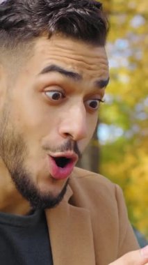 Heyecanlı İspanyol asıllı adam, sonbahar parkında iyi haberler okuyan akıllı telefon okuyor. Mutlu erkek, mutlu internet mesajı alıyor. Telefonla zafer kazanıyor.