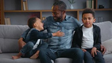 Mutlu Afro-Amerikan ailesi gülüyor kanepede gülüyorlar neşeli babaları ve iki küçük çocukları var çocuklar sarılıyorlar kameraya bakıp eğleniyorlar.