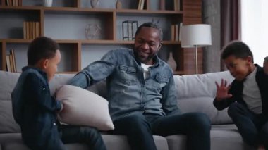 Afro-Amerikan ailesi kaygısız oğulları babalarına yastıklarla vuruyor küçük neşeli yaramaz çocuklar babalarıyla kavga ediyorlar gülüyorlar kanepede eğleniyorlar yastıklarla kavga ediyorlar