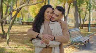 Hispanic couple hugging in autumn park man guy hug beloved girl woman from behind back in autumnal city outdoors embracing enjoy conversation talking together cuddling gently hugging wife with husband