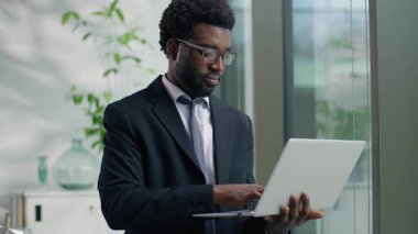 Pencereden dışarı bakan Afro-Amerikalı işadamı modern ofiste düşünceli erkek profesyonel iş adamı olarak çalışırken şirket stratejisi planlayıp gülümsüyor.