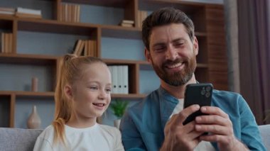 Kafkasyalı mutlu aile telefonlarına bakıp gülen baba-kız gülüşlerine gülüyorlar. Mobil video içerikli komik kız çocukları izliyorlar. Akıllı telefona bakıyorlar.