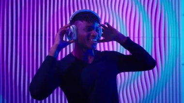 Neşeli duygusal Afrikalı Amerikalı adam fütürist neon klübünde kulaklıkla müzik karışımı dinliyor mutlu DJ erkek adam müzik dinliyor fütürizm alanında gülücük dansı yapıyor eğleniyor