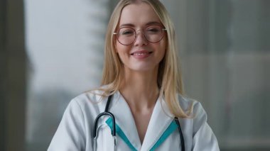 Kafkasyalı mutlu, gülümseyen kadın doktor kalp şeklinde kardiyolog, hemşire bayan kız, klinikte çalışan kameralı pratisyen hekime bakarak kalp hareketi gösteriyor.