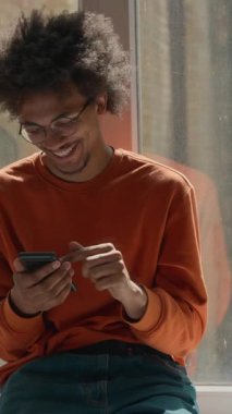 Üniversite koridorunda gülümseyen cep telefonu kullanan Afro-Amerikalı öğrenci. Pencere kenarında dinlenen mutlu adam. Akıllı telefon uygulaması.