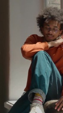 Afro-Amerikalı öğrenci, üniversitenin pencere eşiğinde oturup kedileri okşuyor, kızlar geliyor, erkek, üniversitenin evcil hayvanı, uyukluyor, arkadaş canlısı, üç çok ırklı, kadın öğrenci, gülümsüyor, arkadaşlık çağında.