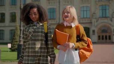 Üniversite kampüsünün dışına çıkan çok ırklı bayan öğrenciler gülümseyen Afrika kökenli beyaz kız arkadaşlar sınıf arkadaşları lise binasını terk edip liseyi tartışıyorlar.