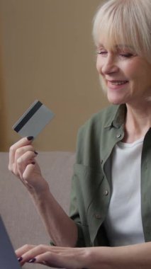 Yaşlı, beyaz, 60 'lı yaşlarda, dizüstü bilgisayarı ve banka kredi kartıyla internetten alışveriş yapan yaşlı bir kadın. Evde gülümseyen, internet alışverişi e-ticaret siparişi veren yaşlı, emekli bir büyükanne.