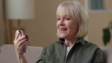 Yaşlı beyaz kadın cep telefonuyla video konuşması yapıyor. Akıllı telefonuyla konferansın keyfini çıkarıyor. Gülümseyen, emekli yaşlı bayan.