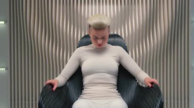 Beyaz modaya uygun bir kadın sandalyeye oturmuş kameraya bakıp gülümsüyor mutlu siber fütüristik kadın kız bakışları neşeli görünüyor içeride oturmuş beyaz fütürizm stüdyo oyun konseptinde poz veriyor.