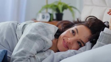 Gülümseyen beyaz kadın, sabah uyandığında neşeyle gülümsüyor. Yeni gün yatak odasında mutlu bir şekilde uzanıyor. Kadın, sıcak, güneşli bir dairede uyukluyor.