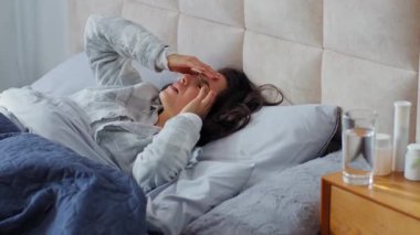 Sağlıksız beyaz kadın, baş ağrısı şikayetiyle üzgün bir şekilde yatakta uyanıyor. Sağlık sorunları var. Kadın, yatak odasında migren gerilimi yaşıyor.