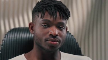 Modern siber fütüristik mekanda gülümseyen Afrikalı Amerikalı adam sandalyeye oturmuş kameraya bakıyor oyuncu adam içeride barışçıl bir şekilde gülümsüyor fütürizmin beyaz stüdyosunun keyfini çıkarıyor.