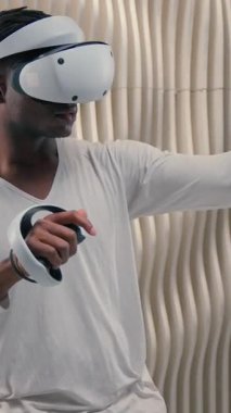 Afro-Amerikan fütürist adam sanal gerçekliği kullanarak VR oyunundan zevk alıyor. Fütürizm stüdyosu oyuncusu erkek oyuncu, eğlence amaçlı sanal ortam için kask tutucu kontrolörleri ile çevrimiçi oynuyor.