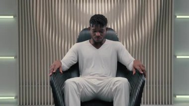 Kendine güvenen Afro-Amerikan fütürist adam koltukta oturmuş kameraman erkek programcıya bakıyor. İçerde oturmuş güven teknolojisi fütürizm odası modern vr uzay ile poz veriyor.