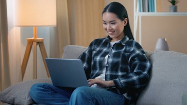 Mutlu gülümseyen Asyalı kadın internette sohbet ediyor video iş bilgisayarları izliyor sosyal medya uygulamasında kanepe genç Japon öğrenci serbest internet alışverişi yapıyor oyun oynuyor.