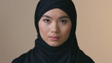 Ciddi bir Asyalı Müslüman kadın portresi, siyah başörtüsü takıyor. Kapalı mekanda yalnız başına kameraya bakıyor. Üzgün kadın başörtüsü kimlik ayrımcılığı yapıyor.