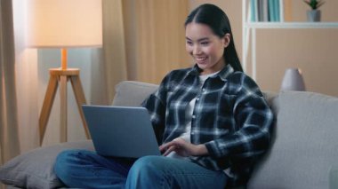 Mutlu gülümseyen Asyalı kadın internette sohbet ediyor video iş bilgisayarları izliyor sosyal medya uygulamasında kanepe genç Japon öğrenci serbest internet alışverişi yapıyor oyun oynuyor.