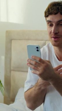 Mutlu Kafkasyalı adam yatak odasında oturmuş akıllı telefon konuşmaları kaydediyor gülümseyen erkek adam internet konferansı sosyal ağlar teknolojisi içerik oluşturma hakkında konuşuyor.