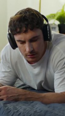 Neşeli Kafkasyalı adam evde yatarken kulaklık takıyor. Akıllı telefon kullanıyor. Dans ediyor, dans ediyor, mutlu erkek.