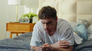 Mutsuz Kafkasyalı adam evde akıllı telefon kullanarak uzanıyor. Kafası karışık, telefon kontrol eden, cep telefonu sorunu olan, kaşlarını çatan, cep telefonu dolandırıcılığı spam uygulama sosyal medya hatasını kaybetti.