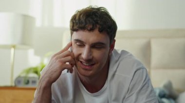 Mutlu, gülen, beyaz bir adam yatak odasında akıllı telefonla konuşuyor. Avrupalı, neşeli, mutlu bir erkek. Mobil telefon görüşmesi yapıyor.