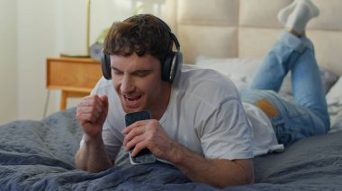 Neşeli Kafkasyalı adam evde yatarken kulaklık takıyor. Akıllı telefon kullanıyor. Dans ediyor, dans ediyor, mutlu erkek.