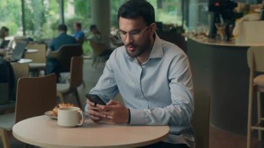 Hintli etnik iş adamı Arap işadamı kafede cep telefonu kullanıyor gülümsüyor kameraman adama bakıyor akıllı telefonlu gülücük atıyor online menü servisi sipariş ediyor teknolojili internete kayıyor