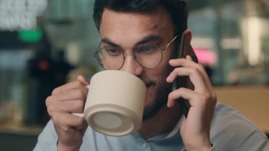 Mutlu Müslüman gülümseyen Hintli işadamı cep telefonuyla konuşuyor, kahve içiyor, kahve içiyor, iş adamı gülüyor, Arap işvereni içkisini içiyor, akıllı telefon iletişiminden memnun.