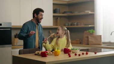 Mutfakta eğlenen mutlu bir aile, neşeli bir beyaz baba, dans eden küçük bir kız çocuğu, elinde mutfak kepçesiyle şarkı söylüyor mikrofonda şarkı söylüyor gibi yapan komik oyun zamanı eğlencesi.