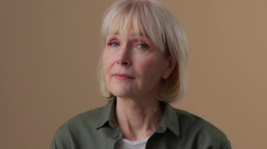 Düşünceli, beyaz, yaşlı bir kadın gülümseyip kameraya bakarken mutlu, mutlu, yaşlı bir bayan 60 'lı yaşlarda, bej renkli stüdyo tebessümü ve sağlıklı dişlerle poz veren mutlu bir büyükanne görüyor.